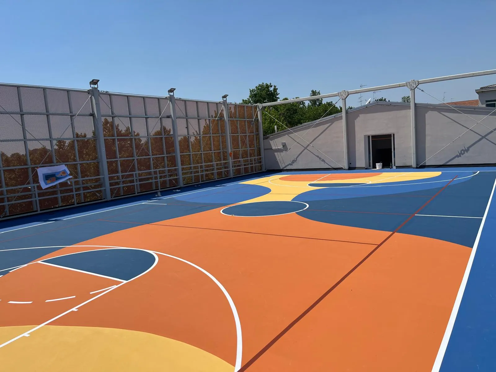 Campo da basket sul tetto della palestra del Liceo Belfiore di Mantova: pavimentazione in resina acrilica Mapecoat TNS pluri-color (arancio, blu, giallo) realizzata da Prima Pavimenti su solaio in calcestruzzo da 15 cm, intervento PNRR 202