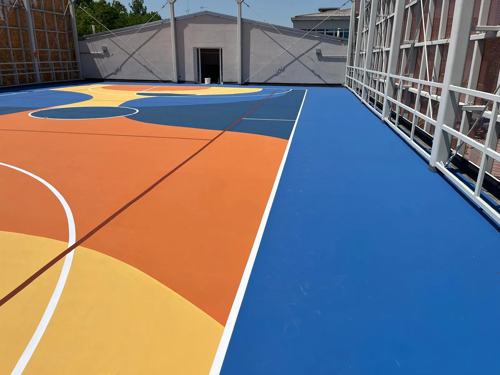 Campo da basket sul tetto della palestra del Liceo Belfiore di Mantova: pavimentazione in resina acrilica Mapecoat TNS pluri-color (arancio, blu, giallo) realizzata da Prima Pavimenti su solaio in calcestruzzo da 15 cm, intervento PNRR 2026